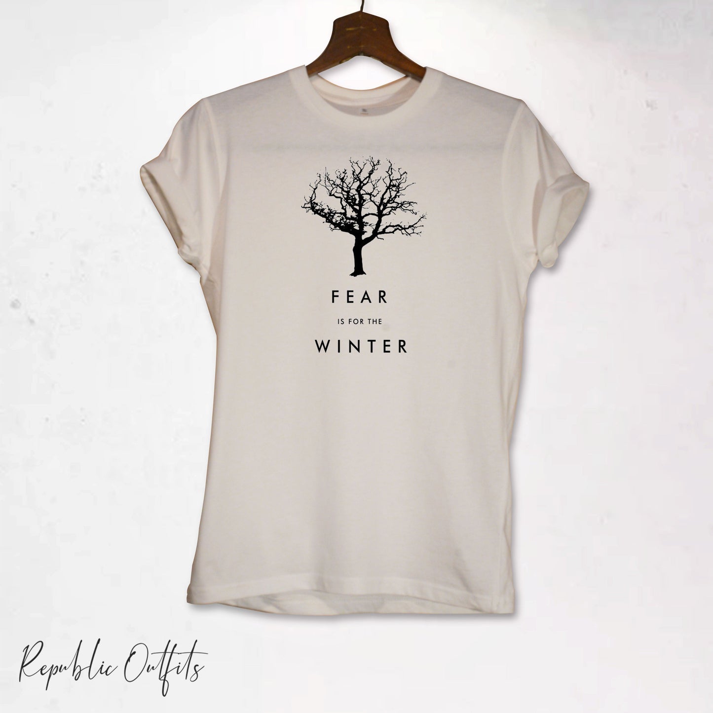 A white crew neck t-shirt with a graphic print of a tree with the text 'FEAR IS FOR THE WINTER' displayed on it, hanging on a hanger against a white background with a black hanging lamp and a brand tag visible.